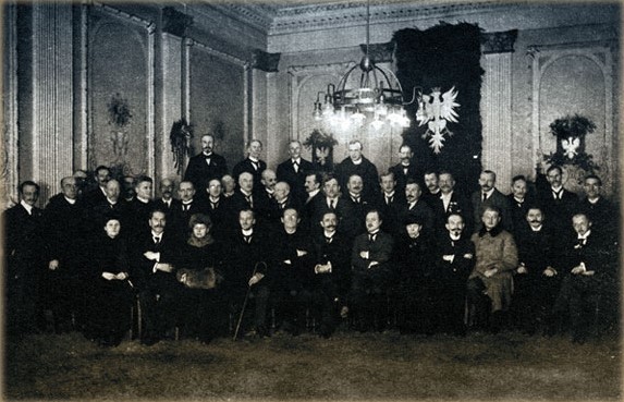 Czarno białe zdjęcie kilkunastu mężczyzn, znajdujących się w sali udekorowanej herbem Wielkopolski. - grafika artykułu